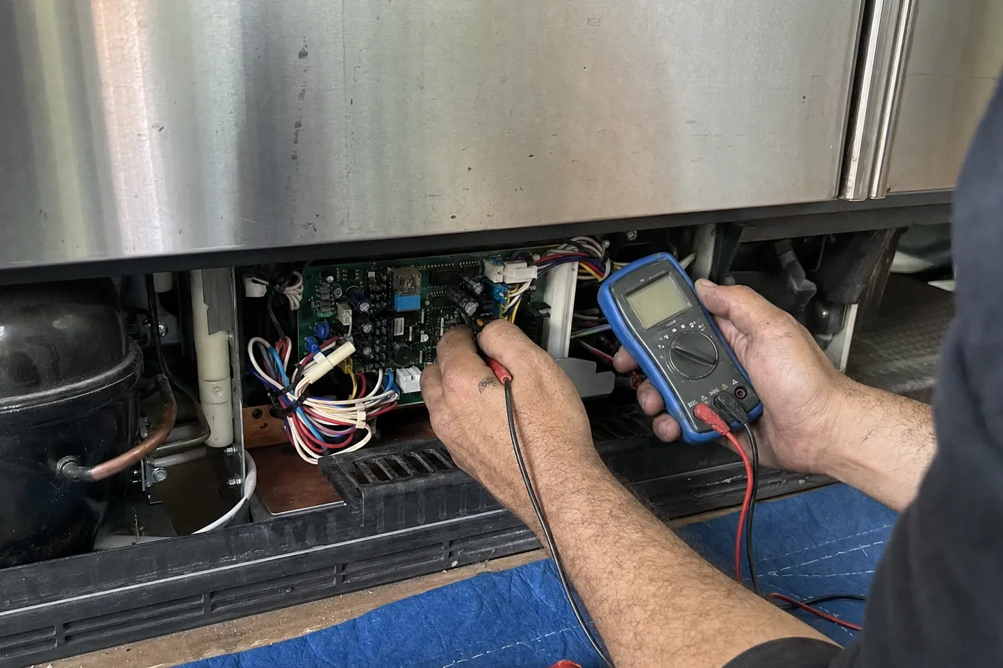 Hands pressing a multimeter probe onto an electronic control board in the lower machine compartment, the wiring loom running off to one side